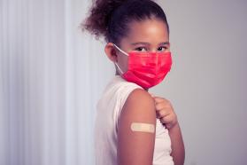 Masked young child holding up sleeve after receiving vaccination