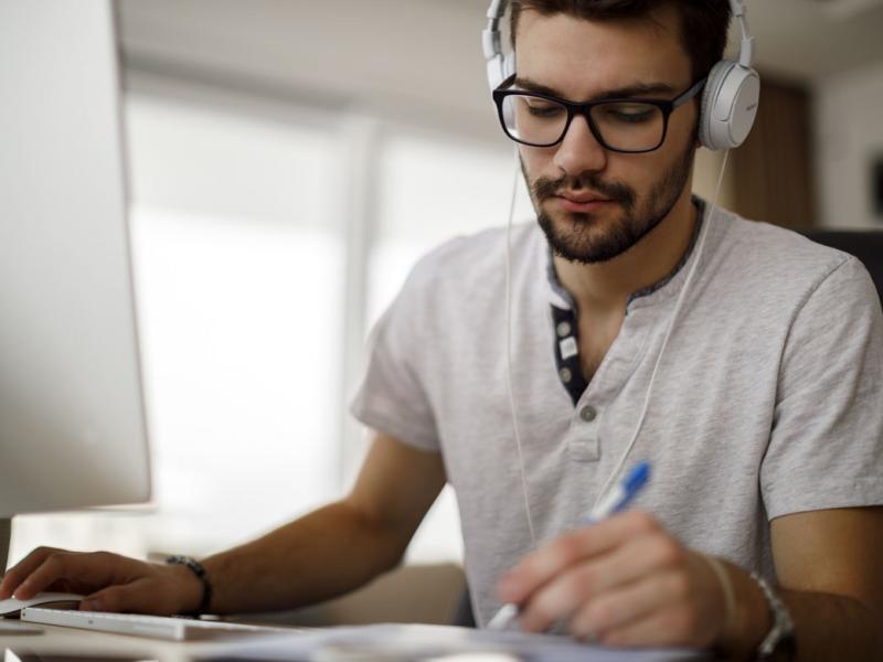 Man wearing headphones working at a computer, taking notes.