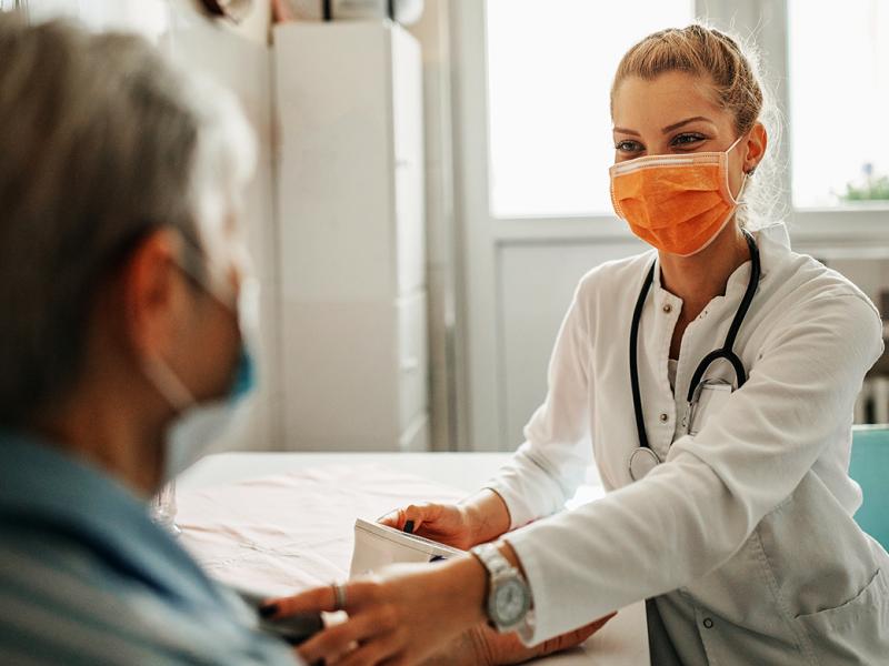 Physician in orange mask sitting down and measuring patient's blood pressure.