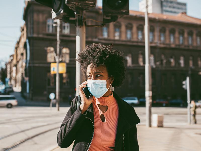 Woman walking down the street wearing a face mask and talking on a cell phone.