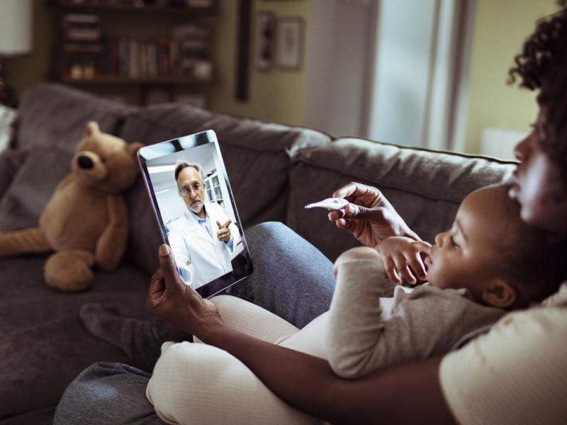 A mother and child sitting on a couch talking to a doctor on a smart pad