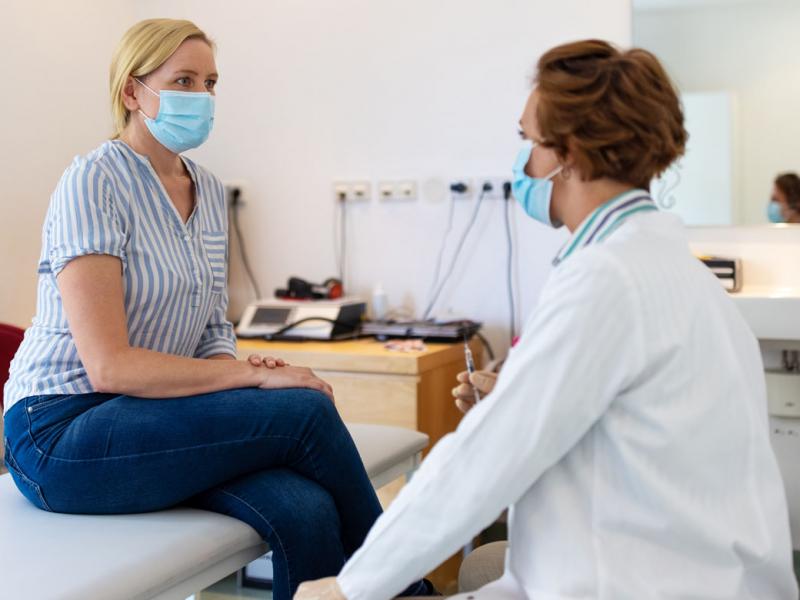 Doctor speaking to a patient sitting on an examination table, both wearing masks.