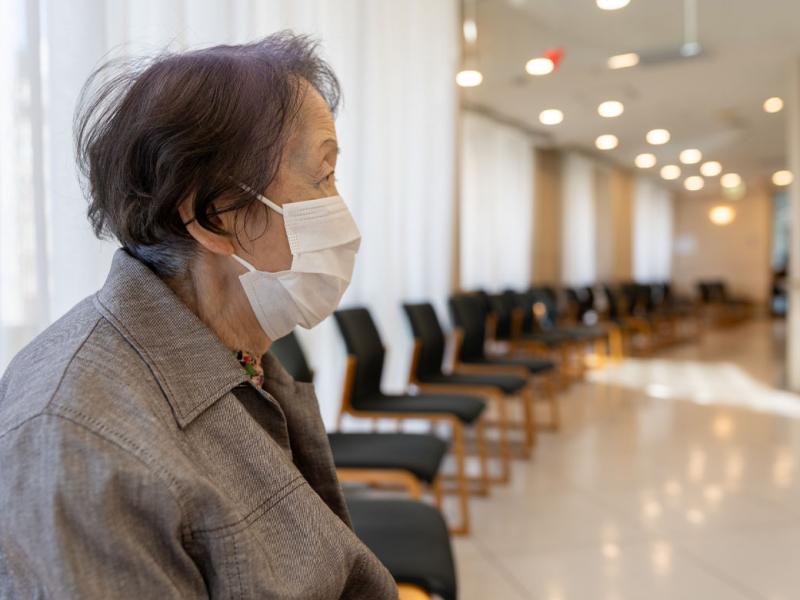 An older woman sitting alone in a hospital waiting room wearing a face mask.