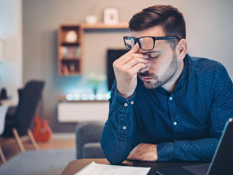 Man sitting at a desk with eyes closed, pinching bridge of his nose with glasses pushed to his forehead.