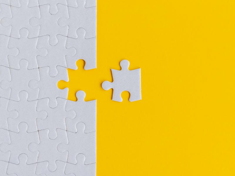 Against a bright yellow background, a close-up of an entirely grey jigsaw puzzle with one piece removed and set to the side of the rest of the puzzle.