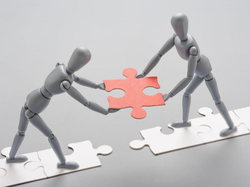 Two grey artist mannequins facing each other, standing on a line of white jigsaw pieces, holding a red jigsaw piece as if to place it to connect the line.