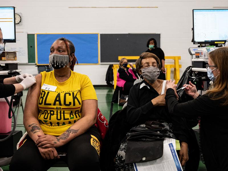 Two women wearing masks and receiving vaccinations at a school or communinity center.