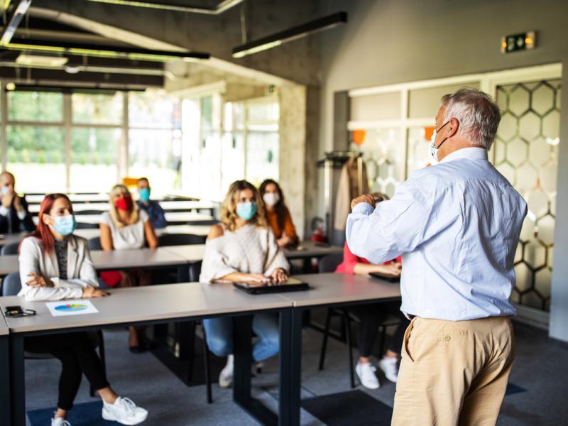 Man making a presentation in front of a group of college age people, all wearing masks