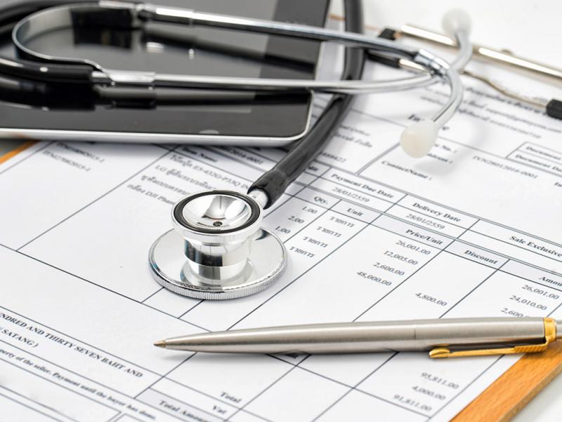 Stethoscope, laptop and pen on top of a medical bill attached to a clipboard