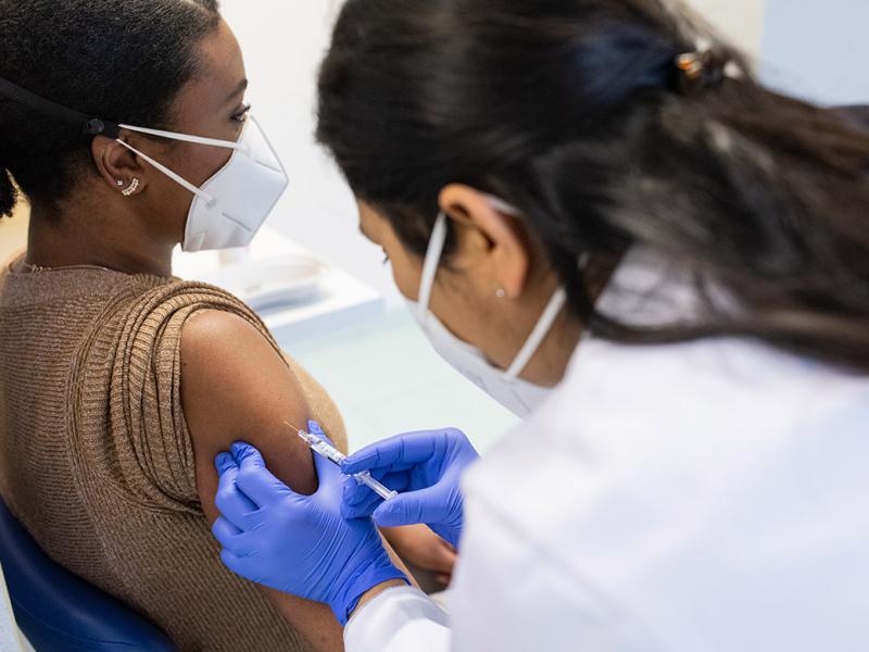 Side shot of a patient wearing a mask and sitting in a chair while a masked health professional gives her a vaccination on her right arm 