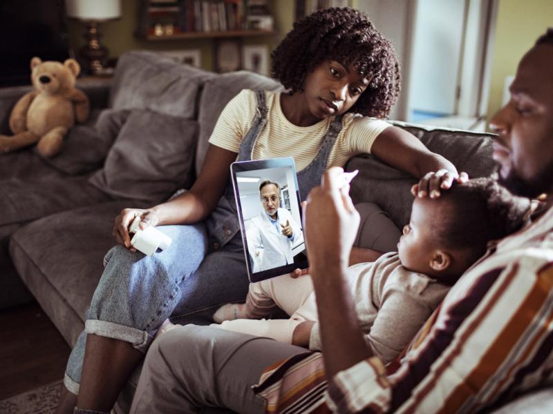 Father and mother sitting on couch with their ill baby having a telehealth call with a physician; the father taking the baby's temperature and the mother feeling the baby's forehead
