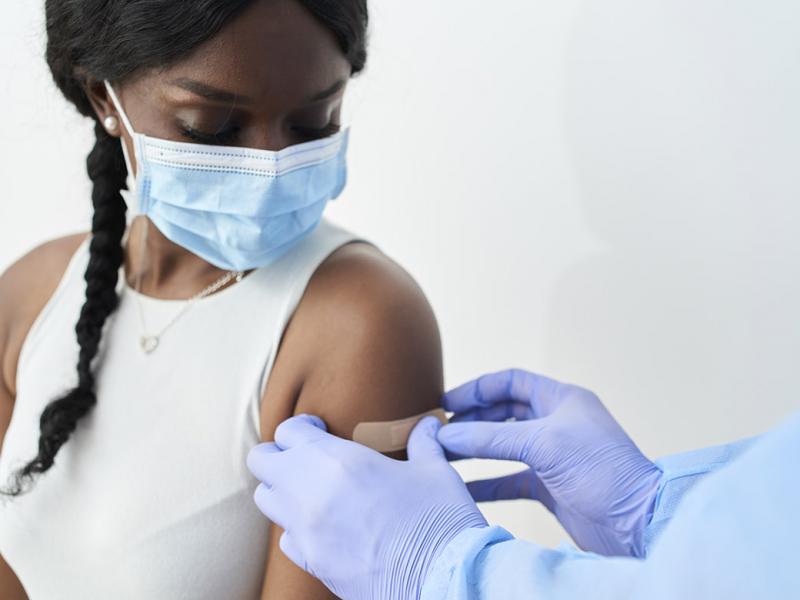 A masked young woman getting an adhesive bandage placed on her left arm after getting a vaccine 