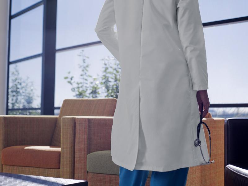 Shot from the back of a physician standing in a hospital waiting area looking out a set of windows, holding a stethoscope in the right hand