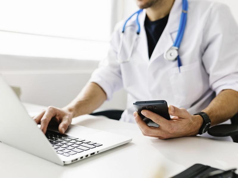 Doctor typing at a laptop with one hand and holding a smartphone in the other hand