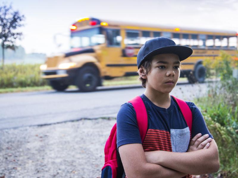Child wearing a backpack with arms crossed, bus in the background