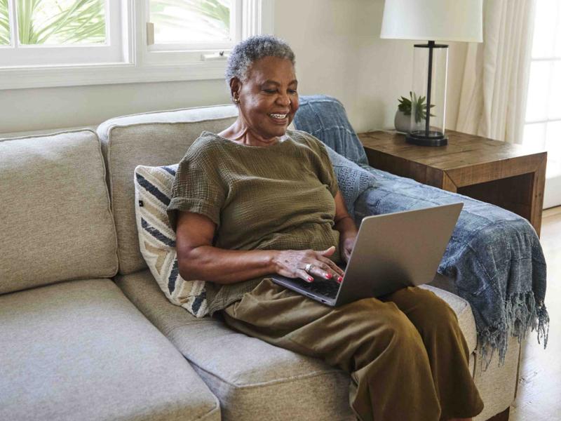 Smiling woman sits on couch while typing on laptop