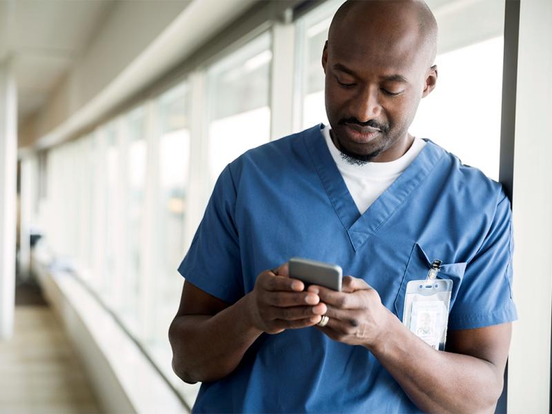 A young physician leans against a wall of windows and looks at his smart phone.