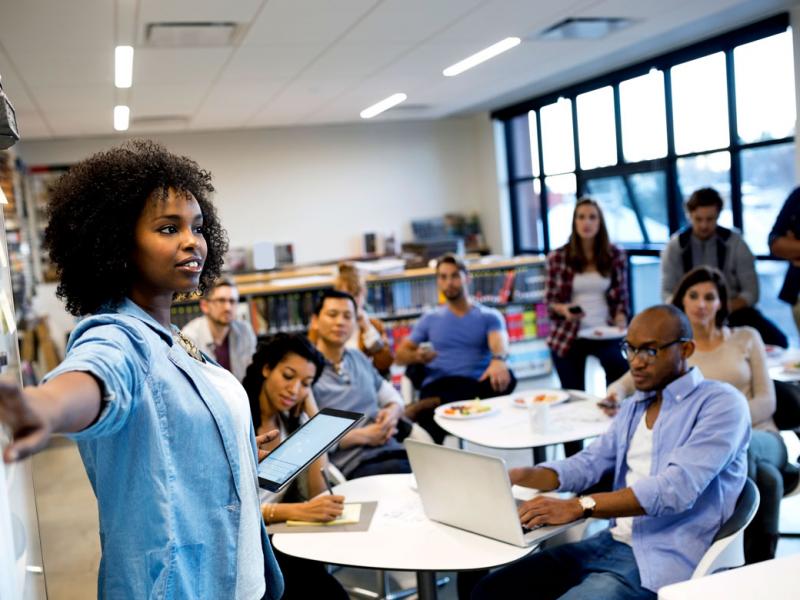 A classroom full of diverse medical students listens to one of their classmates as she explains a procedure using a whiteboard.