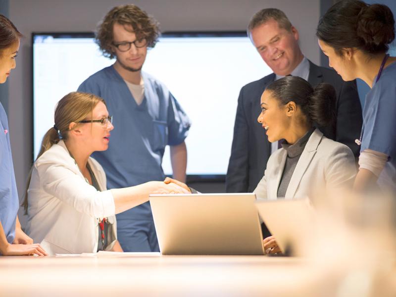 Two physicians shake hands over a laptop while 4 other physicians look on.