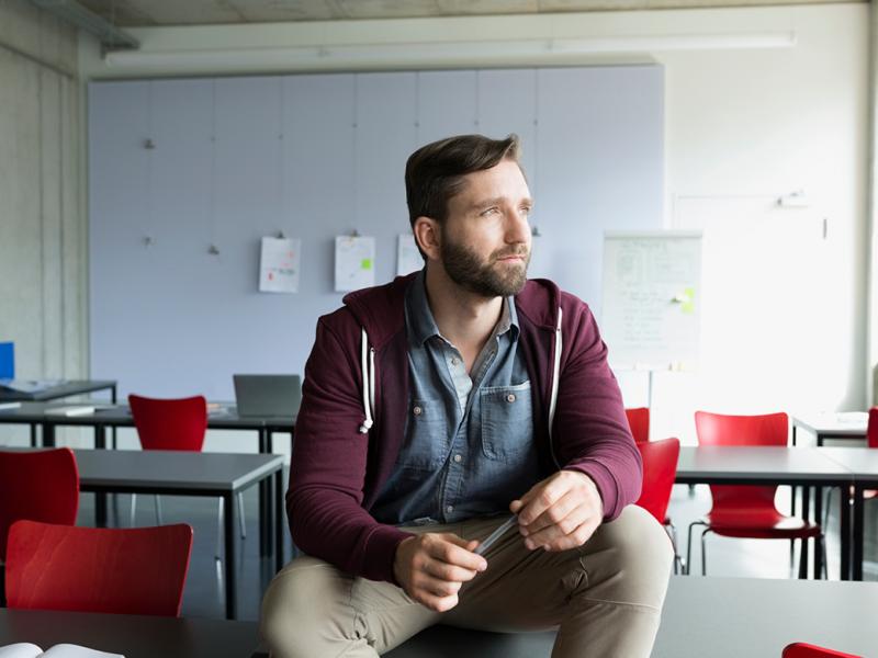 A man sitting on a table in an empty classroom holding a pen and looking out the window.