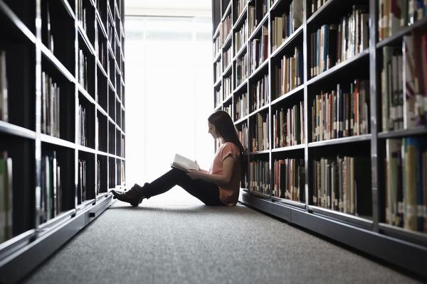 A medical student sits on the floor in a library between aisles of books. 