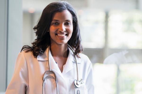 A young physician in a white coat with a stethoscope around her neck smiles and looks at the camera.