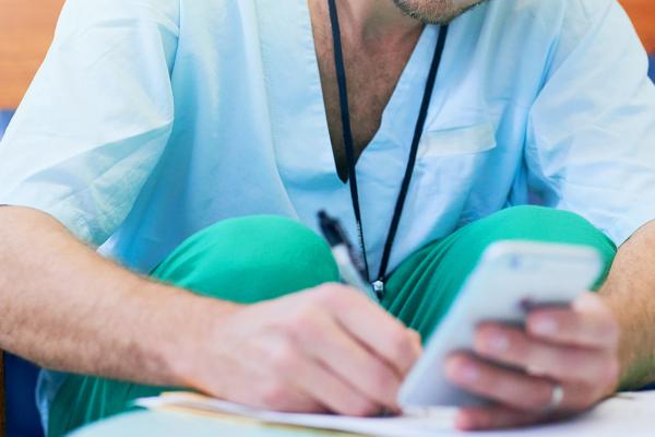 A doctor writing on a notepad while looking at a smartphone.