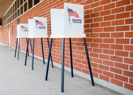 Three punch-tab style voting booths with an American flag and 'VOTE' on their sides, outdoors in front of a brick wall.