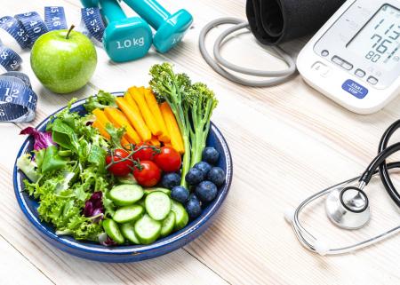 Clockwise, a measuring tape, apple, 1 kg hand weights, a BP monitor, a stethoscope and a plate of fruits and vegetables.