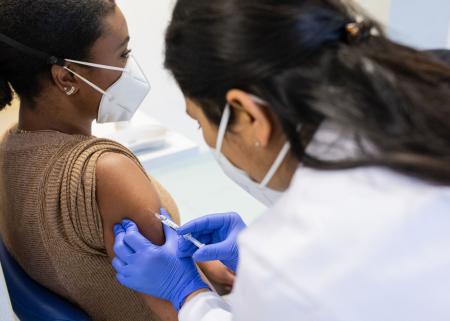A young African-American woman wearing a mask receiving a shot from a health care worker wearing gloves and a mask.
