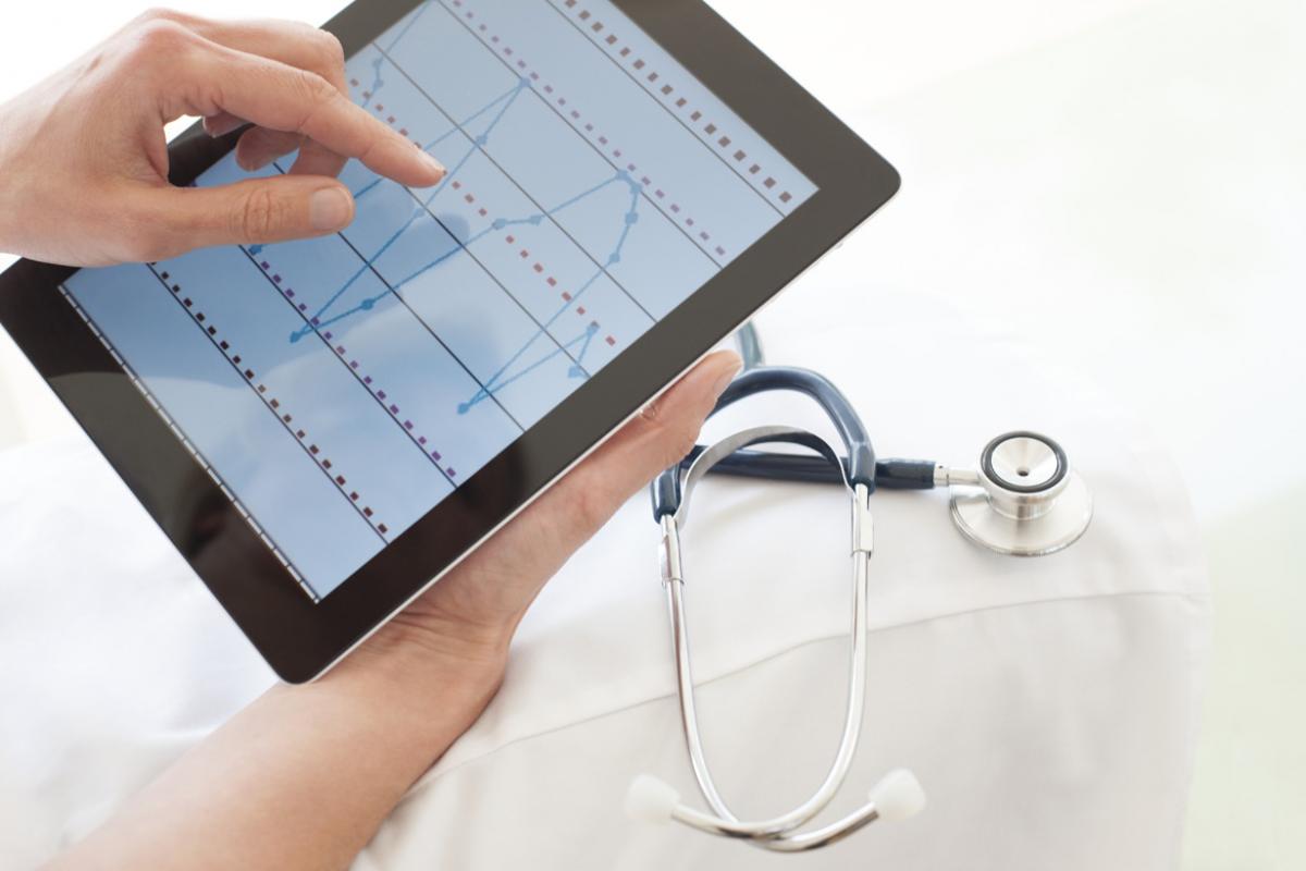 Person holding tablet that shows waves on the screen, next to white coat and stethoscope