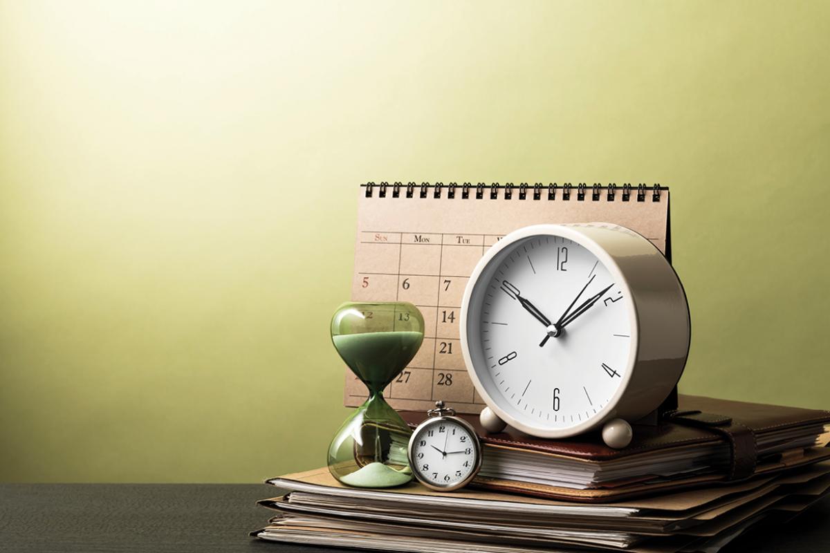 Two clocks, an hourglass, a calendar and an agenda book on top of stack of folders on desk.