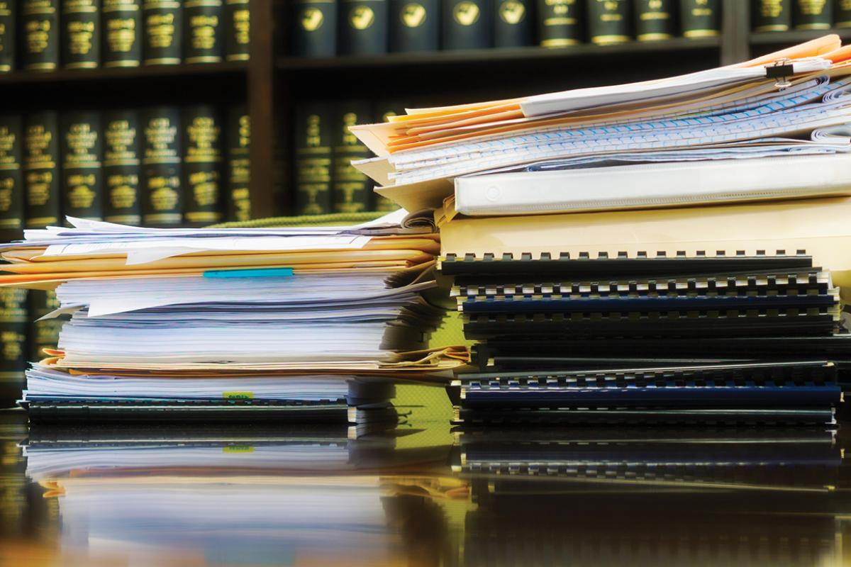 Stack of papers and folders on desk with law books in background.