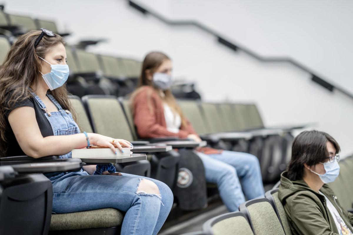 Three students sitting in an auditorium wearing masks and distanced from each other.