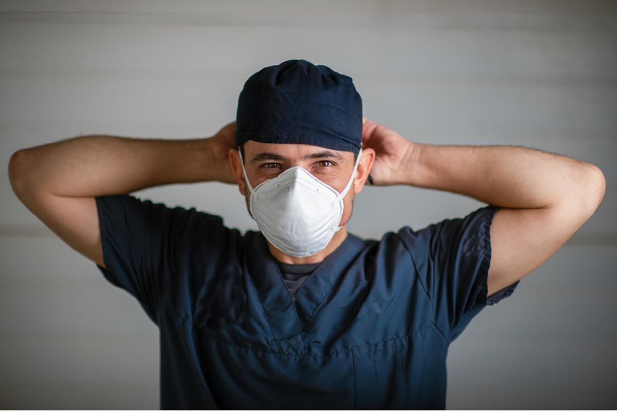 Chest up view of a doctor in scrubs tying on a mask.