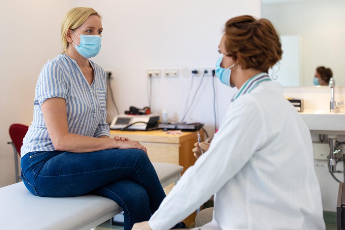Doctor speaking to a patient sitting on an examination table, both wearing masks.