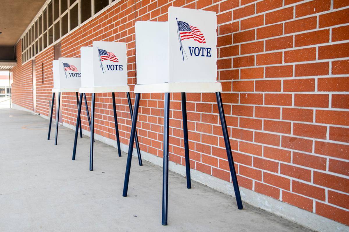 Three punch-tab style voting booths with an American flag and 'VOTE' on their sides, outdoors in front of a brick wall.