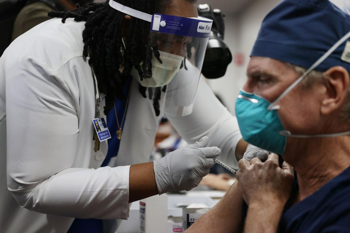 A health care worker in PPE giving a vaccination to a physician in a mask.