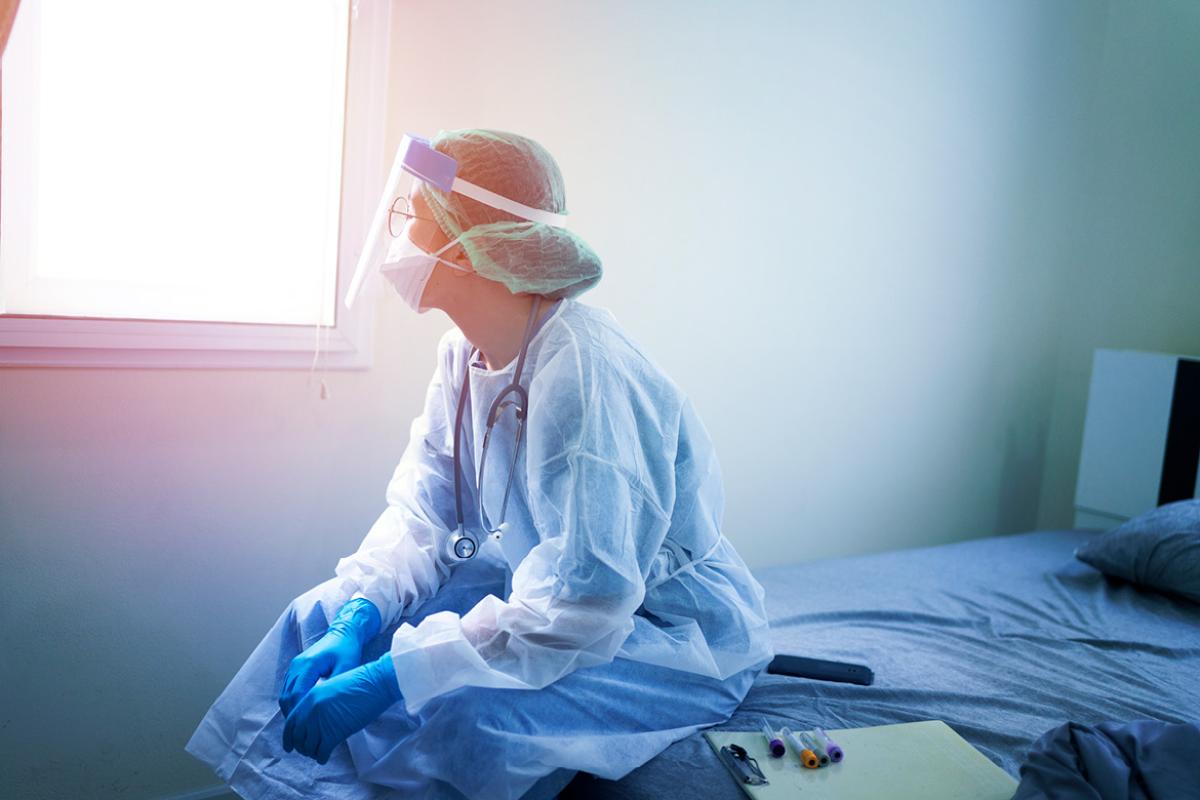 A nurse in full PPE sitting on the edge of a hospital bed looking out of a bright window.