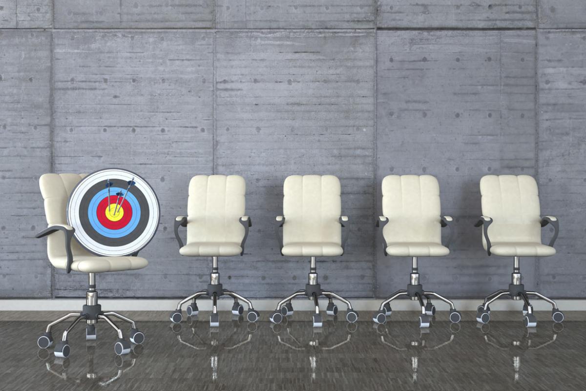 A line of five white office chairs against a grey wall, with the first the first chair holding an archery target pierced by three arrows.