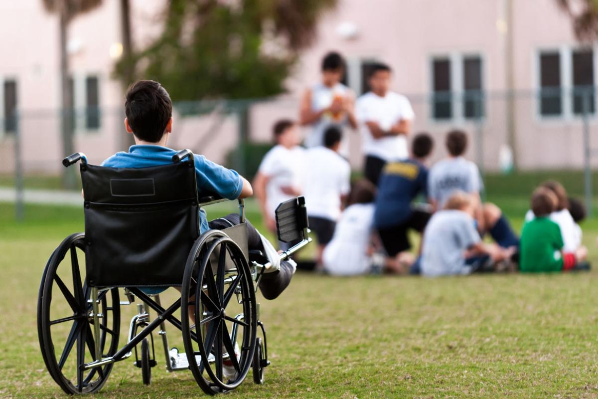A child in a wheelchair in the foreground looking at a group of children playing in the background.