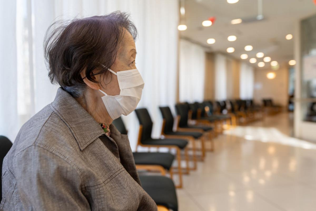 An older woman sitting alone in a hospital waiting room wearing a face mask.