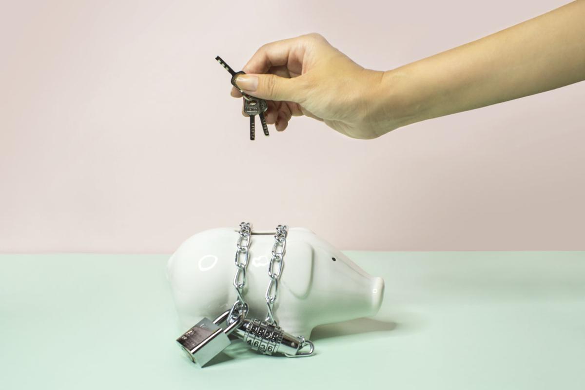 Person holding a key over a chained up piggy bank against a pastel pink and blue background