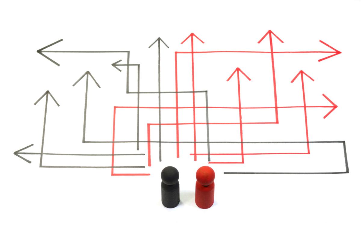 Two wooden block people, one black and one red, standing on a white background facing a number of paths represented by multiple, angular black and red arrows drawn in marker.