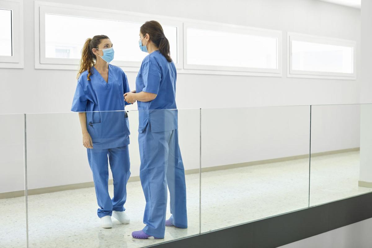 Two physicians in blue scrubs and masks talking in an all white hallway.