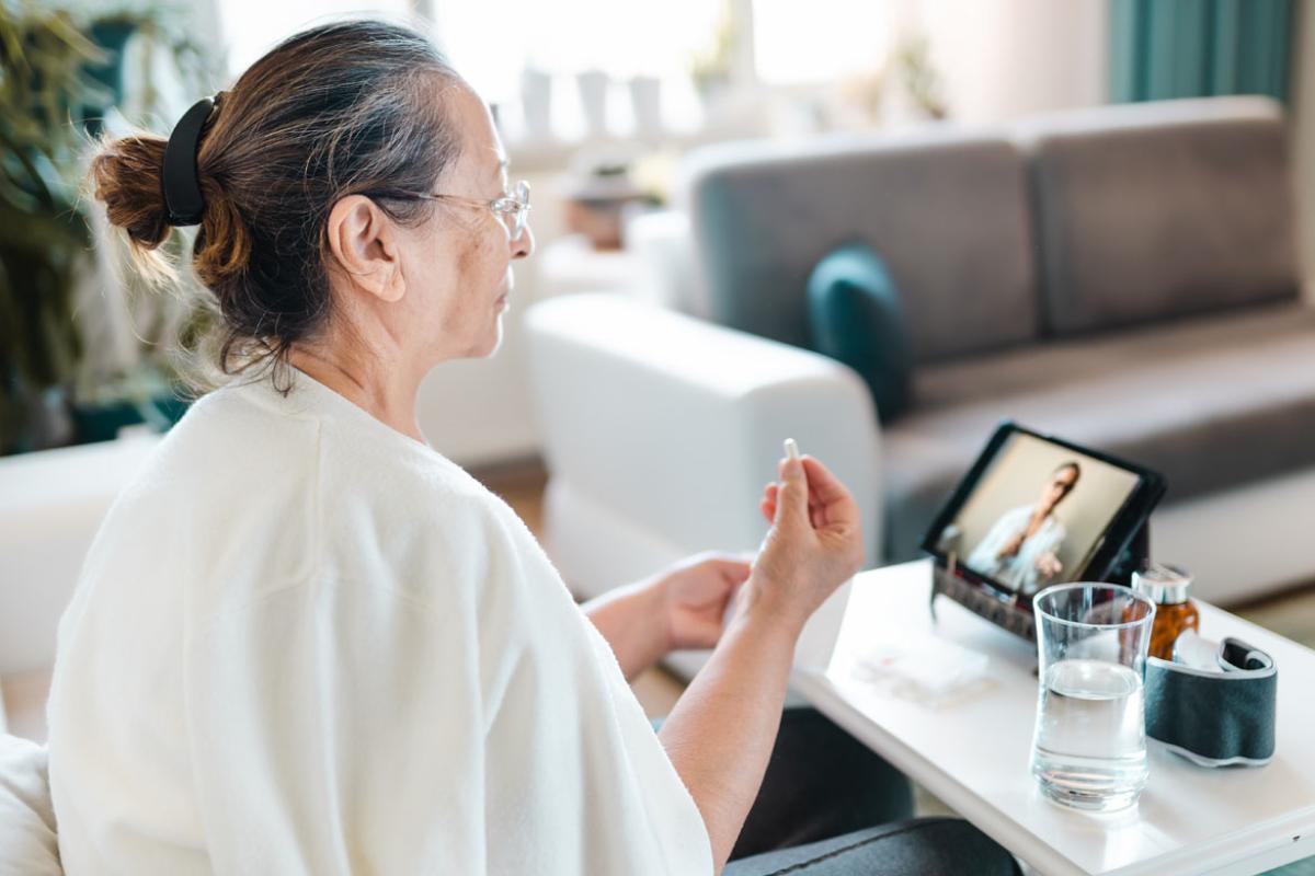 An older woman holding a pill and speaking to a health care worker via teleconference.