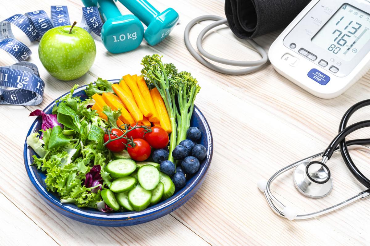 Clockwise, a measuring tape, apple, 1 kg hand weights, a BP monitor, a stethoscope and a plate of fruits and vegetables.
