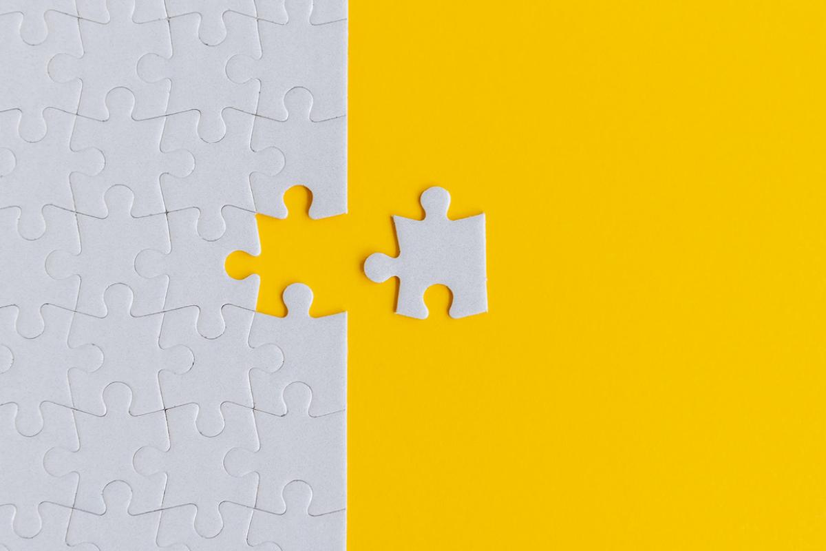 Against a bright yellow background, a close-up of an entirely grey jigsaw puzzle with one piece removed and set to the side of the rest of the puzzle.