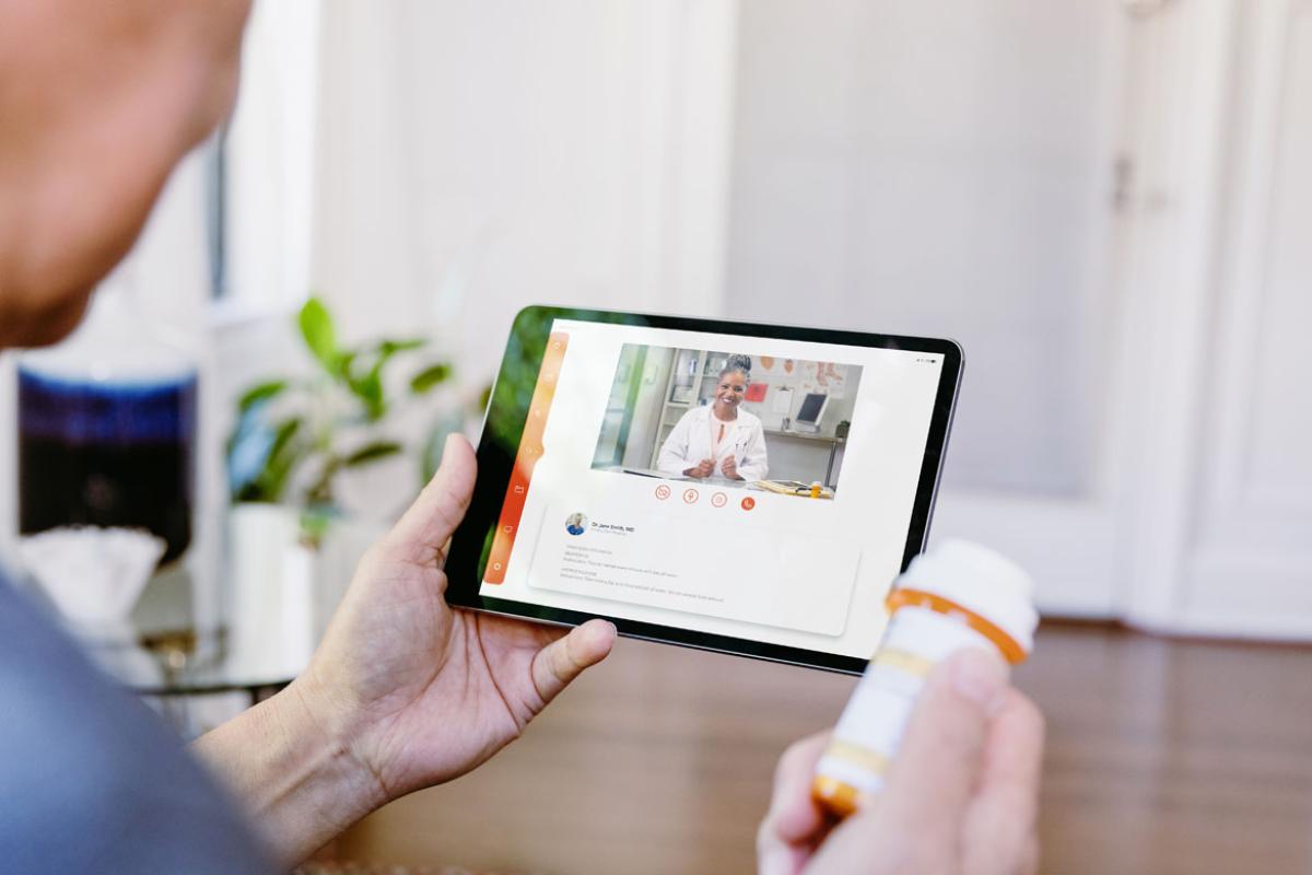 Close-up of a patient's hands, with a prescription bottle in one hand and a smart-tablet with a consulting doctor on the screen.