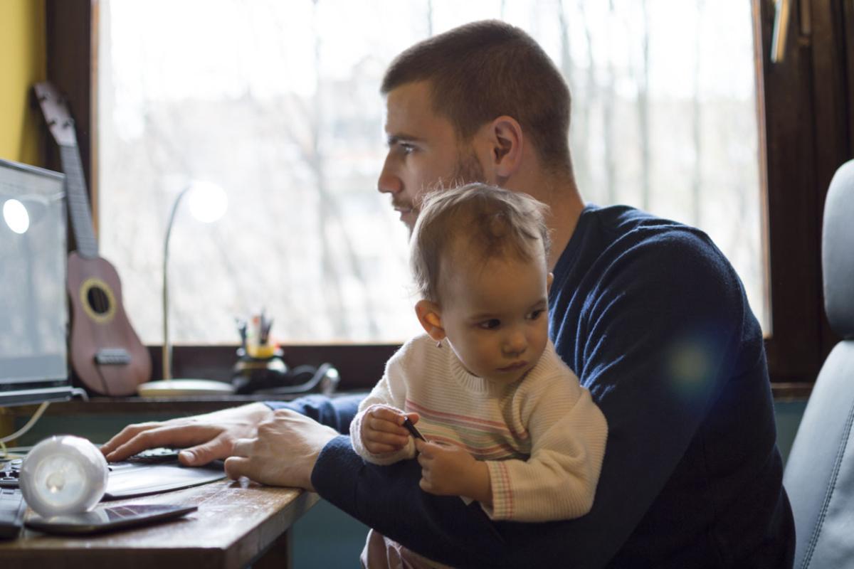 Man sitting at his desk working at his laptop while holding a baby in his lap.
